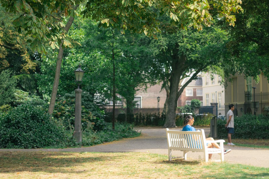 A woman sitting on a white park bench in a shaded communal garden area, reading or resting during a home relocation process, with a young girl standing nearby on a walkway. The garden is surrounded by lush green trees, bushes, and a lamppost, with residential buildings visible in the background. The scene is well-lit, with natural sunlight filtering through the leaves, highlighting the peaceful environment typical of exterior moving or packing logistics. This setting illustrates an outdoor space often used during furniture transport or packing and moving activities, and the presence of the bench and greenery suggests a calm area where relocation staff or homeowners may pause during a removal project, such as organizing or loading belongings, supporting the context found in content about common problems with council parking permits for Holland Park removals.
