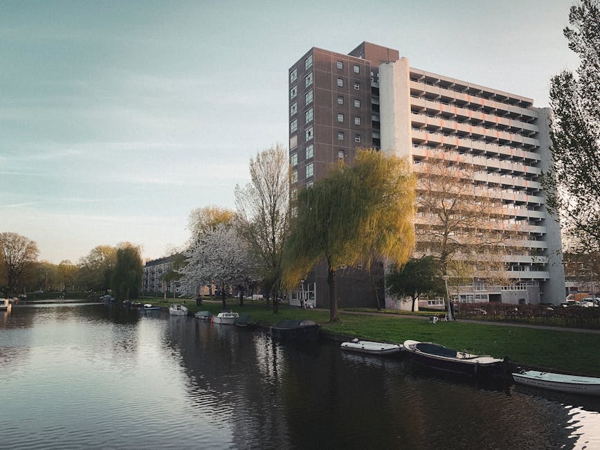 A multi-storey residential building with a white and brown facade is situated beside a canal in an urban park setting. The building features open balconies on each floor and large windows. In the foreground, the canal has several small boats moored along its banks. Trees with blossom and fresh green leaves line the grassy area adjacent to the water, with some trees partially obscuring the lower floors of the building. The scene is illuminated by natural daylight, creating reflections on the water surface. This tranquil environment, with calm waters and greenery, is typical of a peaceful residential area in London. Holland Park Man and Van might use such a location as an example of a house or flat removal environment in the Holland Park district, demonstrating the process of transporting furniture and belongings in a scenic, accessible setting.