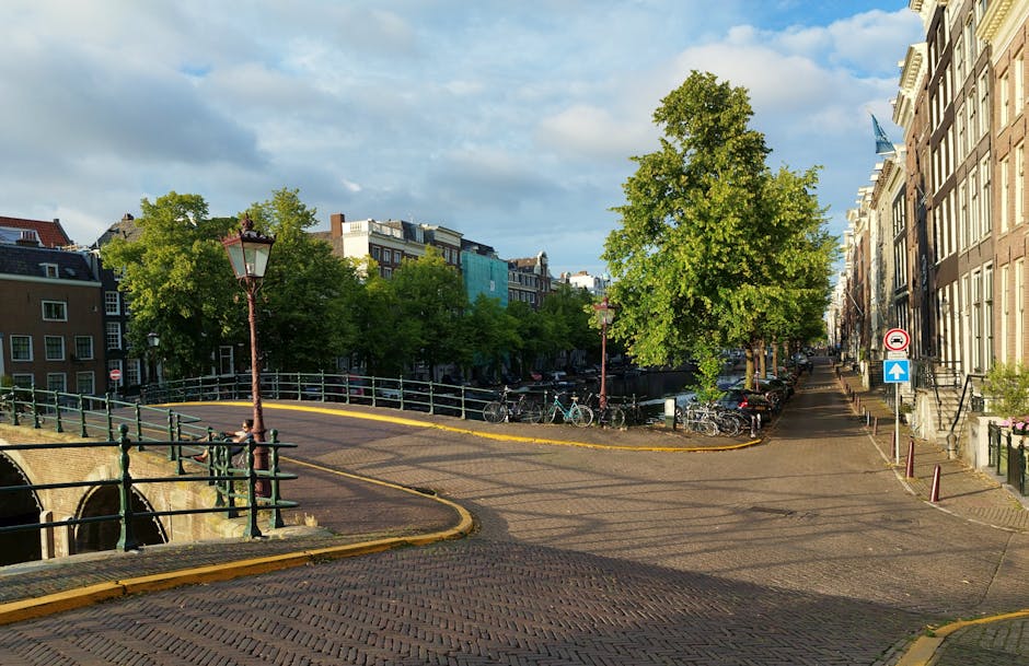 A narrow residential street in Holland Park during daytime, showing a section of the pavement with a parked bicycle rack and several bicycles nearby. There is a small bridge with black metal railing crossing over a canal, with a vintage-style lamp post adjacent to it. The street is lined with multi-storey Victorian-style houses on the right, featuring large windows and ornate facades, and greenery from large leafy trees on the left. A van from Holland Park Man and Van is parked on the right side of the street, with the side door open, indicating a loading process as a cardboard box and a small piece of furniture are being moved or loaded. Visible packing materials such as plastic wrap and blankets are used to protect furniture during the home relocation. Additional boxes and packing materials are inside the van, while some are on the pavement ready for transport. The scene demonstrates a typical setting for furniture transport and packing prior to a house move, reflecting logistical considerations for removals in sheltered urban environments.