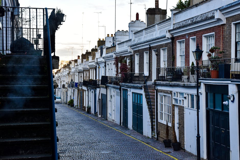 A narrow cobblestone street in Holland Park with historic brick residential buildings on either side, some with white window frames and potted plants along the pavement. Bicycles are parked beside the houses, and a large church or historic building with a tall spire is visible at the end of the street under a clear blue sky. The scene suggests a quiet area suitable for house removals and furniture transport, with potential access considerations for move crews, as seen in the context of home relocation services provided by Holland Park Man and Van.