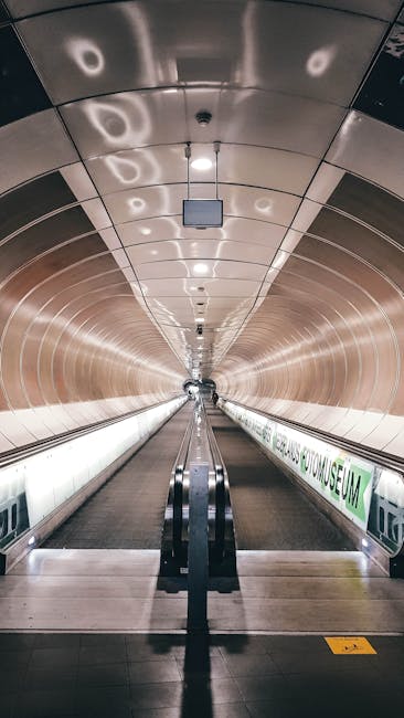 A long underground passageway with a curved, metallic ceiling reflecting the ambient lighting, forming a tunnel-like appearance. The corridor is illuminated by evenly spaced round ceiling lights, creating a well-lit environment. On the right side, a green and white sign displays information about the underground station, indicating it is used for home relocation and moving logistics purposes. The floor is smooth, with a central moving walkway or escalator, and safety barriers and handrails are visible, supporting the loading process for moving furniture or boxes. The environment appears clean and modern, suitable for transporting household items during a house removal or relocation service. The scene is typical of a busy transport hub used by companies like Holland Park Man and Van for coordinating moving operations efficiently, ensuring safe and smooth furniture transport in urban settings.