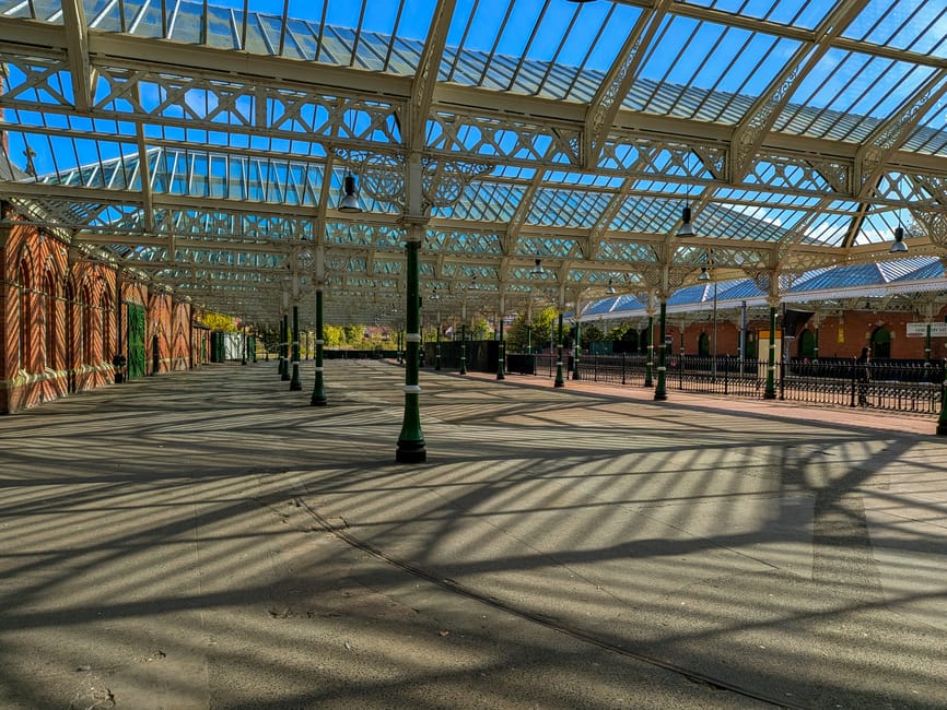 An outdoor area with a large, curved, glass and metal roof structure supported by black metal posts, casting striped shadows on the paved ground beneath. To the left, a brick wall with green doors and windows, decorated with hanging plants, borders the space. On the right, a low black metal fence separates the main area from an adjacent platform with a smaller roofed structure. In the background, trees and a building with yellow walls are visible under a clear blue sky. The scene appears to be part of a transportation or station environment, potentially related to home relocation or moving services, with the roof structure providing shelter for loading or waiting areas, consistent with the services offered by Holland Park Man and Van.