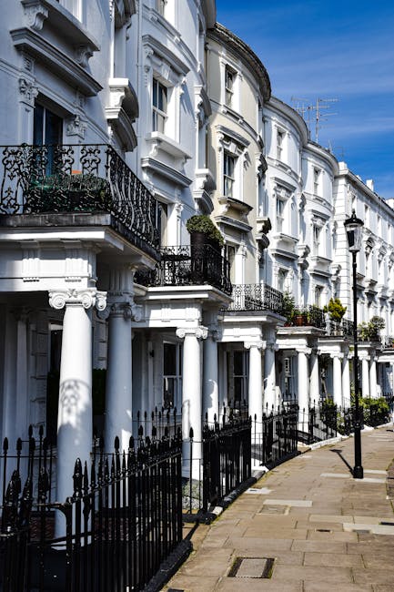 A row of white, Victorian-style terraced houses with ornate architectural details and black wrought iron balconies on the second floor. The buildings feature large sash windows and decorative mouldings, with some small potted plants on balcony railings. The pavement in front of the houses is paved with light-colored stone, and black metal railings separate the properties from the sidewalk. A Holland Park Man and Van vehicle is parked nearby, likely involved in a home relocation or furniture transport process, as part of an ongoing moving or packing project. The scene is illuminated by natural daylight, highlighting the historic charm of the residential street and the potential logistics involved in house removals in the Ilchester Place W11 area.