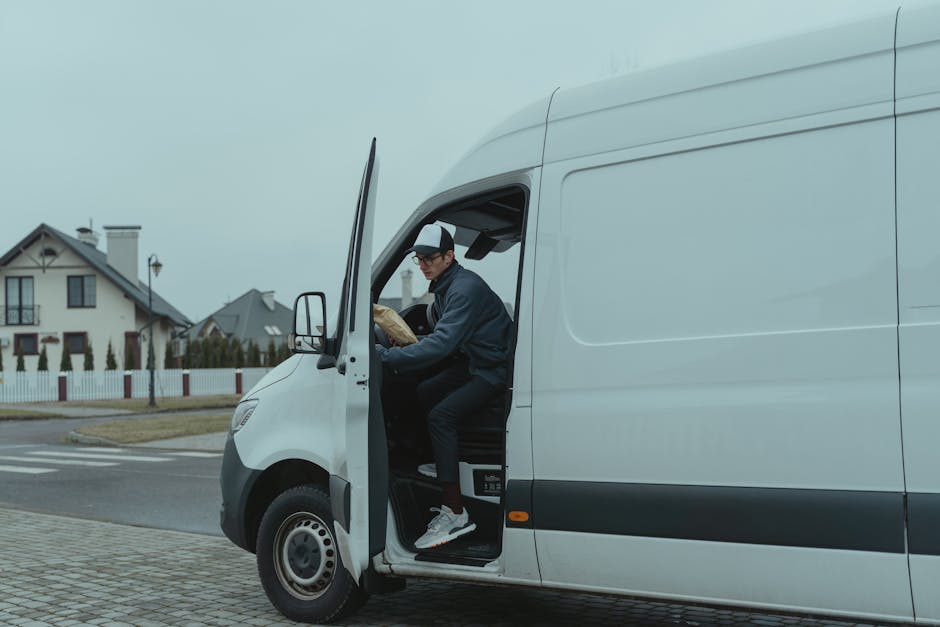 A man dressed in casual dark clothing, wearing a black cap, is sitting in the driver's seat of a white cargo van with the door open. He is holding a brown cardboard box, preparing to load or unload it. The van is parked on a paved street near a residential area with modern houses, white fences, and a lamppost visible in the background. The scene is set during daytime with overcast skies, and the environment appears to be a quiet neighbourhood. The image captures a moment typical of home relocation or furniture transport, illustrating aspects of packing, loading process, and the use of a commercial vehicle for removals, consistent with services offered by Holland Park Man and Van. The focus is on the interaction between the person and the moving equipment, highlighting logistical aspects of a house move.