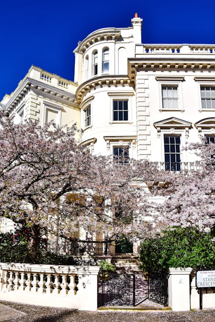 A large white Victorian-style residential building with ornate architectural details, including decorative cornices and rounded bay windows, situated behind a low white brick wall with a wrought iron gate. In front of the property, there are flowering cherry blossom trees with light pink blooms and lush green shrubs, creating a scenic entrance scene. The building is set against a bright blue sky, indicating clear weather. The scene suggests a prestigious area suitable for home relocation or furniture transport, with the property likely being part of a house removal or moving service provided by Holland Park Man and Van, as indicated by the context of house removals and relocation logistics. The overall environment appears calm and well-maintained, suitable for professional packing, loading, and transport activities within an urban setting.