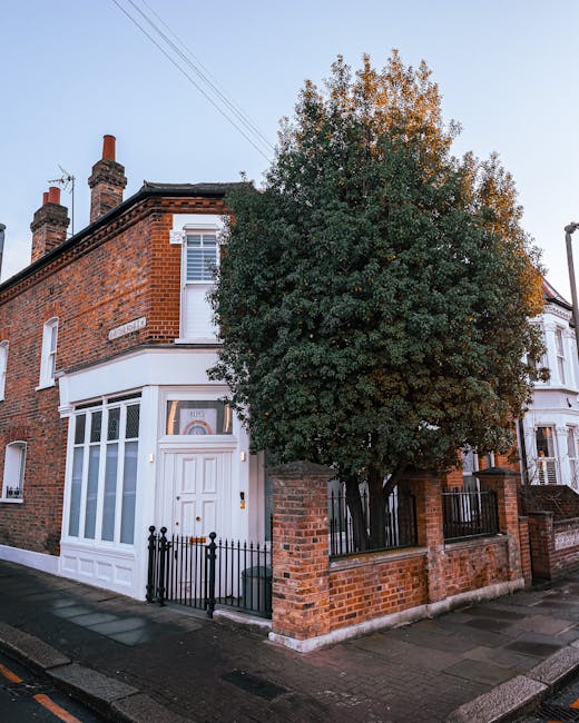 Exterior view of a traditional British semi-detached house on a residential street, featuring a brick facade with white window frames and a front porch. A large, leafy tree partially obscures the front of the property, with the tree situated next to a low brick wall and black metal fence. The pavement in front of the house is concrete, and the sky is clear, indicating daytime. In the context of home relocation and furniture transport, Holland Park Man and Van would coordinate the safe packing and moving of items from this property, which may involve loading furniture, boxed belongings, and appliances into a van for transportation to a new address. This scene reflects the typical environment for house removals in the Notting Hill Gate area, emphasizing the importance of careful logistics and handling during the loading process in a residential setting.