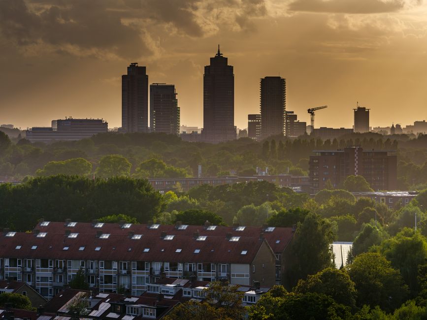 A panoramic view of a city skyline at sunset, with several high-rise buildings, including one with a spire, silhouetted against a sky with scattered clouds. In the foreground, a residential area with 